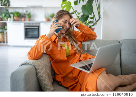 Contented woman uses laptop, talks on smartphone, smiling, laughing, making phone call sits on sofa. Contented woman uses laptop, talks on smartphone, smiling, laughing, making phone call sits on sofa. 119040910