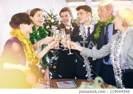 Excited colleagues of different ages with New Year tinsels clinking glasses of champagne during corporate party in office 119044946