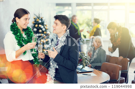 Two joyful male and female coworkers with tinsels and glasses of champagne during corporate New Year party in office Two joyful male and female coworkers with tinsels and glasses of champagne during corporate New Year party in office 119044998