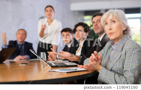 Concentrated young adult old office workers sitting around the table and looking at screen in office Concentrated young adult old office workers sitting around the table and looking at screen in office 119045100