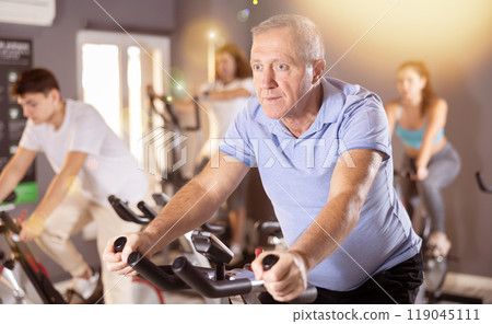 Portrait of mature man taking indoor cycling class at fitness center, doing cardio riding bike 119045111