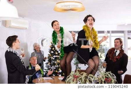 Happy office workers celebrate new year in office - women dance on table, men look at them with delight Happy office workers celebrate new year in office - women dance on table, men look at them with delight 119045240