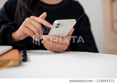 A close-up of an Asian woman in a black sweater using her smartphone at a table indoors. 119045373