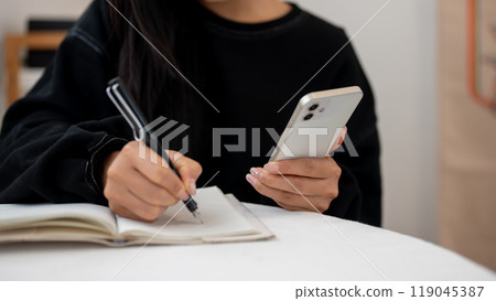 A close-up of a woman writing in a book and using her smartphone while sitting at a table indoors. 119045387
