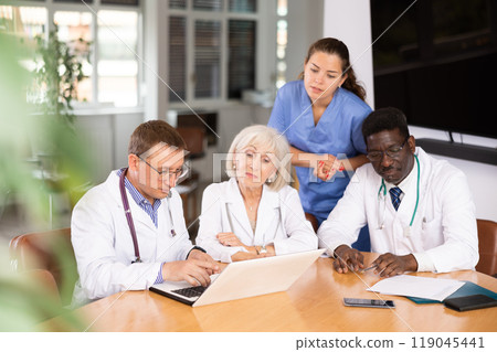 Doctor with colleagues sitting at table, reading new of law on laptop and discussing information Doctor with colleagues sitting at table, reading new of law on laptop and discussing information 119045441