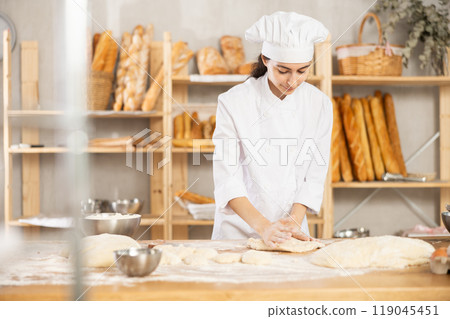 Young woman works as baker, mold baguette from dough, creates long loafs from dough. Young woman works as baker, mold baguette from dough, creates long loafs from dough. 119045451