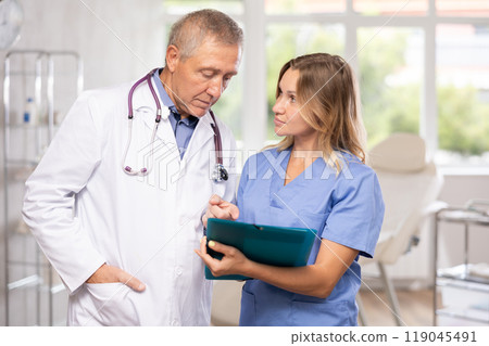 Middle-aged nurse and mature doctor discussing diagnosis standing in cabinet Middle-aged nurse and mature doctor discussing diagnosis standing in cabinet 119045491