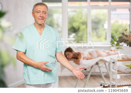 Positive old masseur posing against background of therapy room 119045689