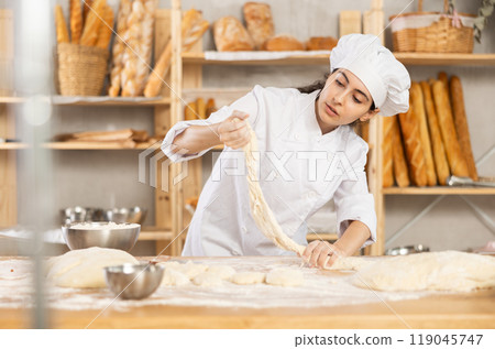 Young woman works as baker, mold baguette from dough, creates long loafs from dough. Young woman works as baker, mold baguette from dough, creates long loafs from dough. 119045747