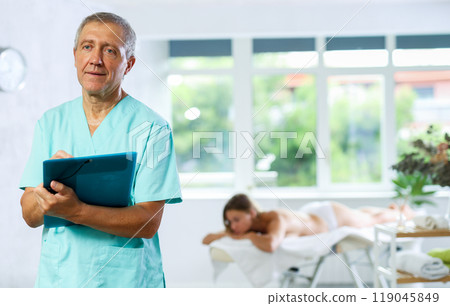 Positive old masseur posing with folder in hand in the therapy room 119045849