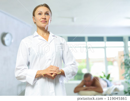 Positive middle-aged masseuse posing against background of therapy room Positive middle-aged masseuse posing against background of therapy room 119045861