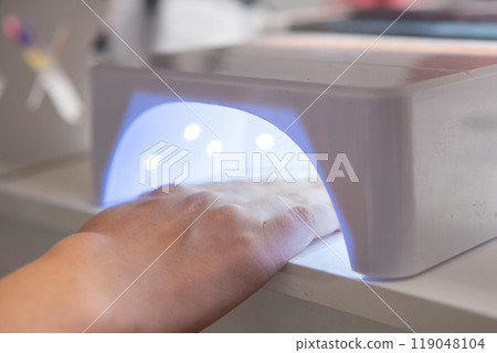 Womans hand is placed under a UV lamp for curing gel nail polish after a manicure. Womans hand is placed under a UV lamp for curing gel nail polish after a manicure. 119048104