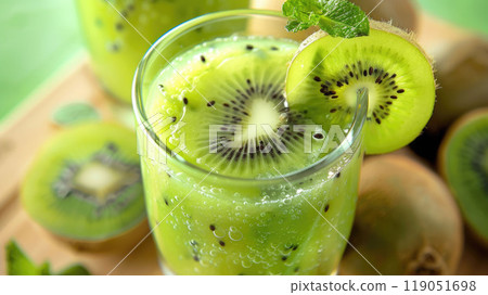 Close-up of a refreshing kiwi smoothie in a clear glass with kiwi slices placed on the rim. The drink is surrounded by fresh kiwi fruits and a few mint leaves for garnish. 119051698