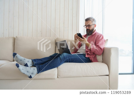 Man with notebook and smartphone sitting on sofa, online banking, internetbanking at home throught website and app. Paying with smartphone app. Man with notebook and smartphone sitting on sofa, online banking, internetbanking at home throught website and app. Paying with smartphone app. 119053289