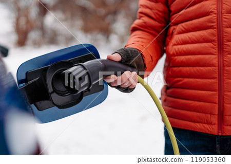 Man charging electric car during cold snowy day. Close up of man putting charger in charging port. Charging and driving electric vehicles during winter season. 119053360