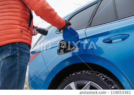 Man charging electric car during cold snowy day. Close up of man putting charger in charging port. Charging and driving electric vehicles during winter season. 119053361