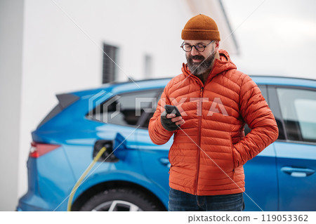 Man charging electric car during cold snowy day, using electric vehicle charging app, checking battery life, energy consumption on smart phone. Charging and driving electric vehicles during winter 119053362