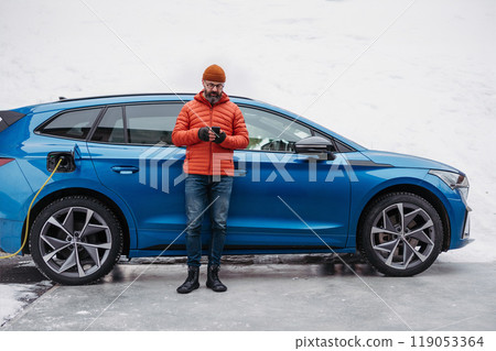 Man charging electric car during cold snowy day, using electric vehicle charging app, checking battery life, energy consumption on smart phone. Charging and driving electric vehicles during winter 119053364