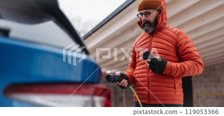 Man charging electric car during cold snowy day. Close up of man putting charger in charging port. Charging and driving electric vehicles during winter season. Man charging electric car during cold snowy day. Close up of man putting charger in charging port. Charging and driving electric vehicles during winter season. 119053366