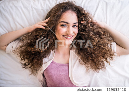 Portrait of beautiful young woman with a curly hair lying on her back on bed. Top view shot. Portrait of beautiful young woman with a curly hair lying on her back on bed. Top view shot. 119053428