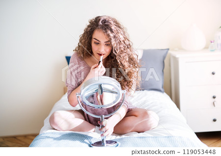Young woman sitting on bed, applying red lipstick to her lips, looking into a round makeup mirror. Young woman sitting on bed, applying red lipstick to her lips, looking into a round makeup mirror. 119053448
