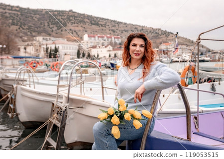 Woman holds yellow tulips in harbor with boats docked in the background., overcast day, yellow sweater, mountains 119053515