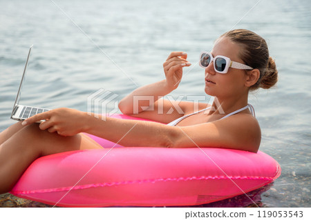 A woman is sitting in a pink inflatable raft on a lake, holding a laptop. Concept of relaxation and leisure, as the woman enjoys her time in the water while working on her laptop. A woman is sitting in a pink inflatable raft on a lake, holding a laptop. Concept of relaxation and leisure, as the woman enjoys her time in the water while working on her laptop. 119053543
