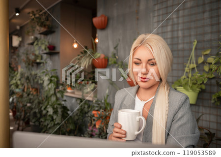 A blonde woman is sitting at a table with a laptop and a white coffee cup. She is wearing a gray jacket and has red lipstick on. The scene suggests a casual and relaxed atmosphere. A blonde woman is sitting at a table with a laptop and a white coffee cup. She is wearing a gray jacket and has red lipstick on. The scene suggests a casual and relaxed atmosphere. 119053589