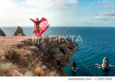 A woman in a red dress stands on a rocky cliff overlooking the ocean. The scene is serene and peaceful, with the woman's flowing dress adding a sense of grace and beauty to the landscape. 119053640
