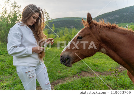 A woman is standing next to a brown horse, looking at it. The woman is wearing a white shirt and white pants. The horse is eating grass. 119053709