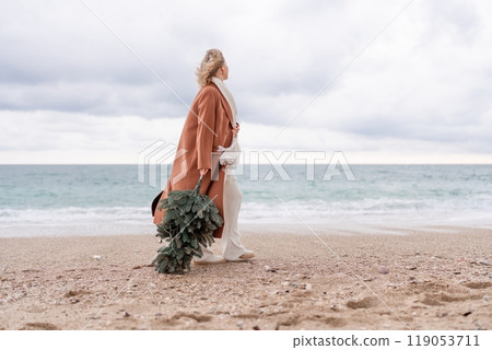 Blond woman Christmas tree sea. Christmas portrait of a happy woman walking along the beach and holding a Christmas tree in her hands. She is wearing a brown coat and a white suit. 119053711