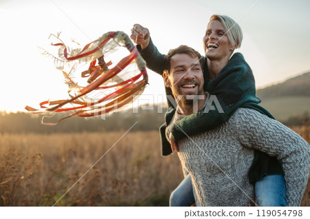 Couple in love on a walk in autumn forest. Husband piggycarrying wife on back, holding wreath with ribbons. 119054798