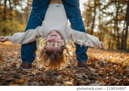 Father holding little girl by legs heand down, playing during walk in autumn forest. Dad and girl having fun outdoors. 119054897