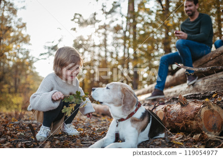 Family on walk resting, sitting on logs in the middle autumn forest. Little girl playing with older dog. 119054977
