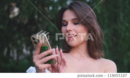 Carefree beautiful woman holding mobile phone, looking at mobile phone screen, using smartphone apps, posing outdoors. 119055187