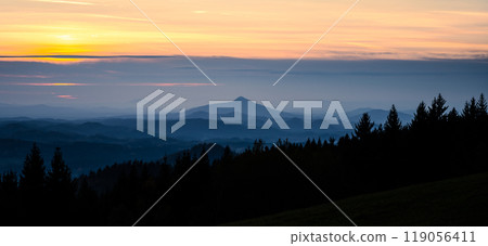 A serene silhouette of hilly terrain unfolds as the sun sets behind Ralsko Mountain in Czechia. The landscape showcases layers of mountains and forests in tranquil evening light. 119056411