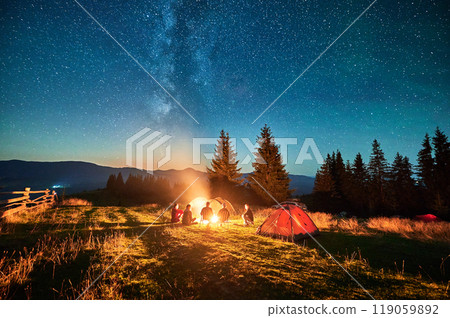 Group of friends hikers resting around campfire, surrounded by illuminated tents under stunning starry sky. Milky Way stretches above, creating magical atmosphere in peaceful mountain wilderness. 119059892