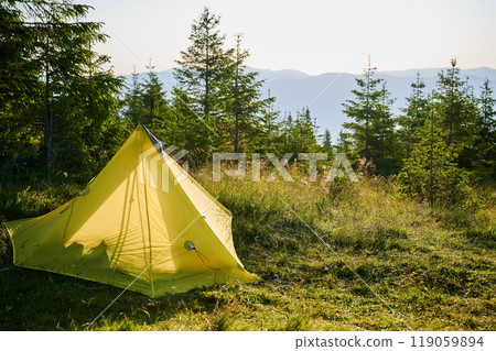 Orange tourist tent on grassy hillside under sunset sky. Surrounded by young pine trees. Stunning view of distant mountains and creates peaceful and picturesque camping spot in the morning. 119059894