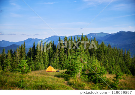 Bright yellow tourist tent on grassy hillside, surrounded by lush green pine trees. Breathtaking view of distant blue mountains under clear sky, creating idyllic and serene camping spot. 119059897