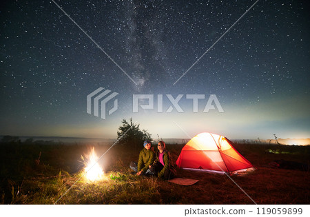 Couple travelers sitting near illuminated camp tent under beautiful sky with stars. Night starry sky over grassy hill with tourist tent, bonfire and hikers. Concept of hiking and night camping. 119059899