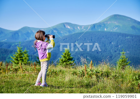 Young girl stands in grassy meadow, looking through binoculars, wearing yellow and purple sweatshirt. Behind her, rolling green hills and mountains stretch into distance under clear blue sky. 119059901