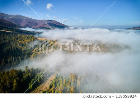 Stunning landscape with dense forests and rolling hills partially shrouded in soft, ethereal fog. Vibrant autumn colors of trees contrast beautifully with clear blue sky and distant mountain peaks. Stunning landscape with dense forests and rolling hills partially shrouded in soft, ethereal fog. Vibrant autumn colors of trees contrast beautifully with clear blue sky and distant mountain peaks. 119059924