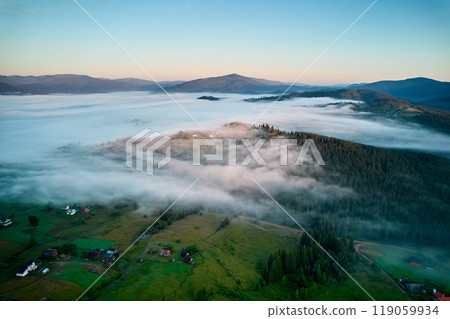 Aerial view of misty morning in mountains, with small village nestled among trees. Fog gently blankets landscape, creating serene and picturesque scene, as sun begins to rise in the background. 119059934