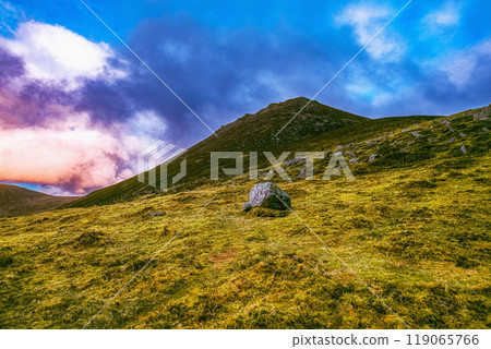A mountain range with multiple peaks in the distance is partially obscured by clouds. The foreground features a grassy field with scattered rocks. A mountain range with multiple peaks in the distance is partially obscured by clouds. The foreground features a grassy field with scattered rocks. 119065766
