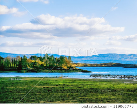 A serene lakeside view showcases vibrant autumn trees along the shore under a bright blue sky with fluffy clouds, creating a peaceful natural setting. A serene lakeside view showcases vibrant autumn trees along the shore under a bright blue sky with fluffy clouds, creating a peaceful natural setting. 119065773