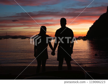 The silhouetted couple stands close together at the beach, enjoying a romantic moment while the sun sets. The silhouetted couple stands close together at the beach, enjoying a romantic moment while the sun sets. 119066223