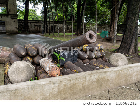 An old cannon rests atop a pile of mortar shells, all arranged in a concrete planter in a park setting. 119066282