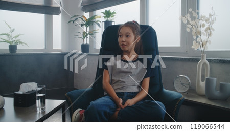 Asian kid sits in chair, talks to female child psychologist during therapy 119066544
