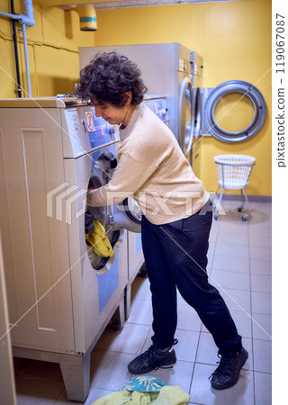 young plus size woman loads clothes into a washing machine in a laundry room in the basement of a student dormitory young plus size woman loads clothes into a washing machine in a laundry room in the basement of a student dormitory 119067087