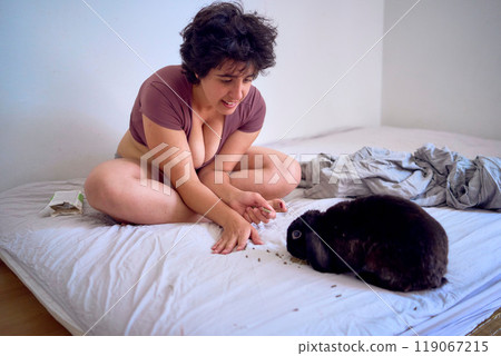 young plus size woman in home clothes playing with her pet rabbit on bed in student dormitory 119067215